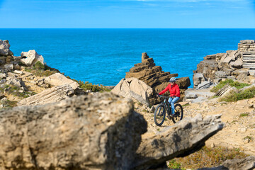 Brave senior woman riding her electric mountain bike on the rocky cliffs of Peniche at the western atlantic coast of Portugal - 789462088