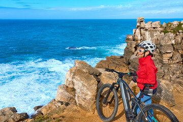 Brave senior woman riding her electric mountain bike on the rocky cliffs of Peniche at the western...
