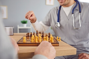 Close up cropped photo of a nurse man playing chess game with a senior elderly person sitting at the table in clinic or in retirement home. Caregiver, healthcare and medical concept.