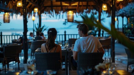 A couple enjoying their evening at an outdoor restaurant with the sea in view, sitting on dark gray rattan chairs and black tables. - obrazy, fototapety, plakaty
