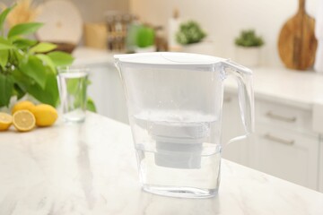 Water filter jug, glass and lemons on white marble table in kitchen
