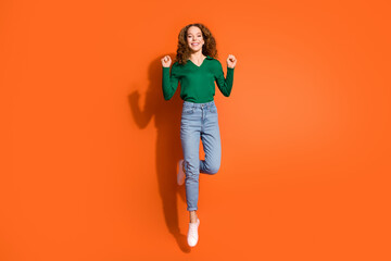 Full length photo of lucky funky lady dressed green shirt jumping high rising fists isolated orange color background