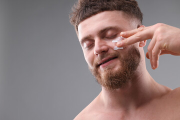 Obraz premium Handsome man applying moisturizing cream onto his face on grey background, space for text