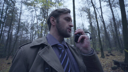 Serious police detective using a walkie-talkie while working at a crime scene in a deep forest 