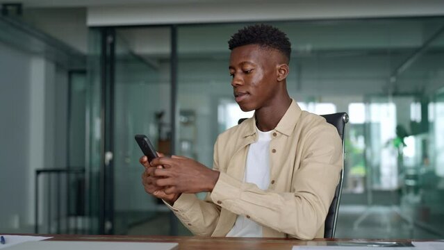 Smiling busy professional African business man sitting in office holding mobile cellphone. Young happy businessman entrepreneur using smartphone looking at cell phone technology device at work.