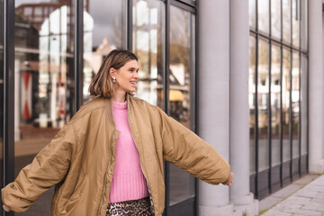 Fototapeta premium Happy blonde short hair woman wear bomber jacket walking in the street and smiling to camera. Stylish girl in fashion outfit walking over the city. What a beautiful day out. Girl turn around.