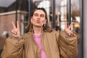 Close up cool blonde short hair woman in bomber and pink sweater shows peace sign, blows kiss and...