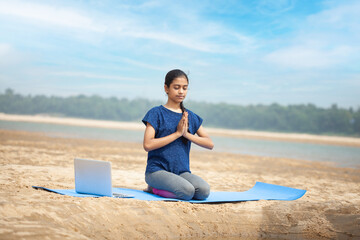 yoga and meditation, indian girl practicing kneeling prayer yoga pose or vajrasana using laptop technology in the calm sea beach, Pranamasana