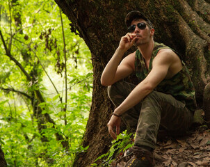 Guerrilla. Portrait of a caucasian adult with strong arms wearing a camouflaged uniform and sunglasses, sitting in the ground while taking a smoke in the tropical forest.	
