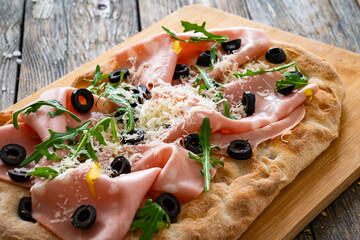 Roman Pinsa with mortadella, parmesan cheese, black olives and arugula on wooden table
