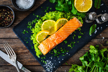 Smoked salmon with lemon and greens on black stony plate on wooden background
