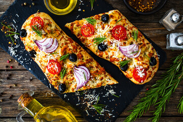 Focaccia - baked sandwich with black olives, tomatoes, onion and rosemary on wooden background

