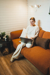 Smiling woman sitting on sofa at home and talking on phone, working on laptop online, freelancer modern lifestyle. - obrazy, fototapety, plakaty