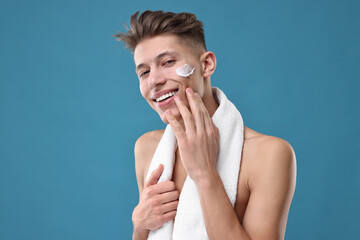 Handsome man applying moisturizing cream onto his face on blue background