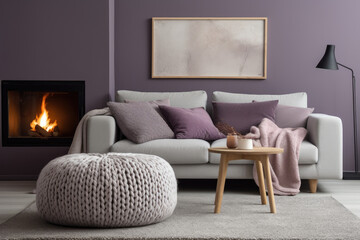 Fototapeta premium Knitted pouf near grey sofas with purple pillows. Fireplace against grey wall with poster frame. Scandinavian home interior design of modern living room