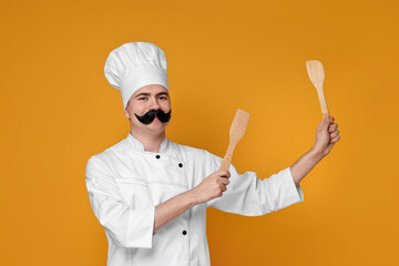 Portrait of happy confectioner with funny artificial moustache holding wooden spatulas on orange...