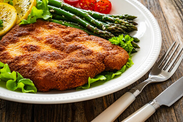 Crispy breaded fried pork chop, green asparagus and fresh vegetables on wooden table
