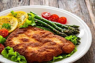Crispy breaded fried pork chop, green asparagus and fresh vegetables on wooden table
