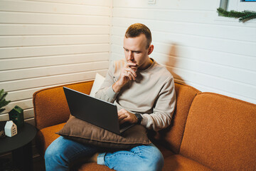 A handsome man in home clothes sits on a sofa and works on a laptop. Remote freelance work.