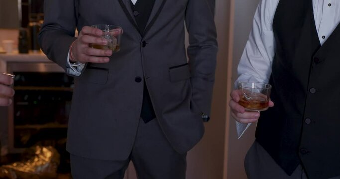Two men in suits, one holding a glass of liquor, stand in a casually elegant room. Their attire contrasts with the laid-back atmosphere, evoking a sense of refined relaxation
