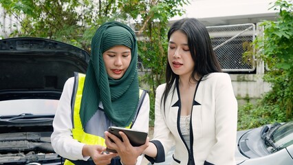 Smiling veiled woman and woman in blazer look at tablet, outdoors, men with car in background....
