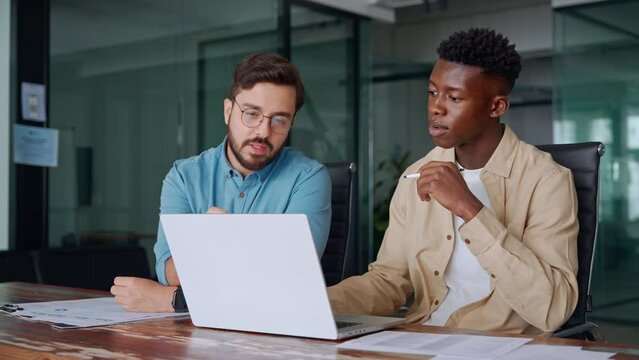 Two busy diverse male employees using laptop computer talking in office. Professional young business men workers team having conversation working on project sharing ideas sitting at work desk.