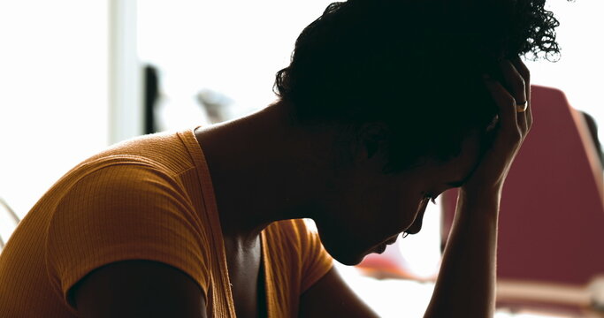 Young African American woman struggles with depression at home in quiet despair feeling overwhelmed by stress and sorrow putting hands on side temples feeling headache