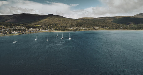 Scotland's sailboats ocean seascape aerial view in coastal water of Brodick Bay. Ships and boats near shore with homes, cottages, roads at greenery valleys. Scottish cityscape scenery shot