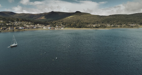 Atlantic ocean gulf, sailboats aerial zooming shot in Brodick Bay. Scottish landscape of port town. Houses, cottages, resort at shore of gulf against greenery lands and mountains view