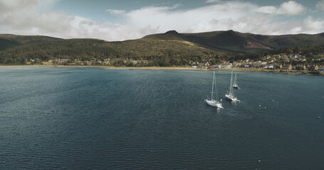 Atlantic ocean gulf, sailboats aerial zooming shot in Brodick Bay. Scottish landscape of port town. Houses, cottages, resort at shore of gulf against greenery lands and mountains view