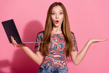 Portrait of impressed teen with long hair wear shirt hold laptop palm presenting object empty space isolated on pink color background