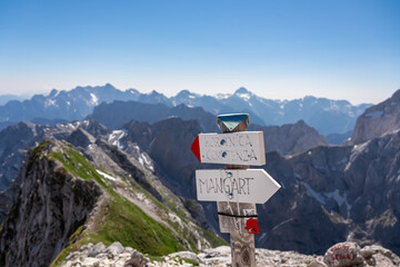 Path mark indicaring hiking trail to mount Mangart (Mangrt), border Italy Slovenia. Scenic view of majestic mountain peaks of Julian Alps on blue sky summer day. Climb rugged extreme alpine terrain - obrazy, fototapety, plakaty