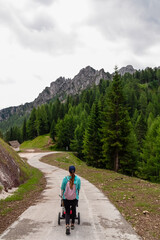 Woman walking with baby carriage along scenic hiking trail on Monte Lussari in Camporosso, Friuli Venezia Giulia, Italy. Looking at majestic mountain ridges of Julian Alps. Wanderlust family concept