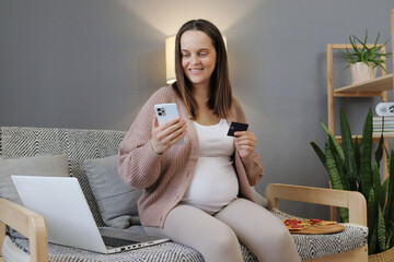 Smiling young pregnant woman shopping on internet from home using banking app or service on smartphone and credit card buying baby clothes online making virtual payment order