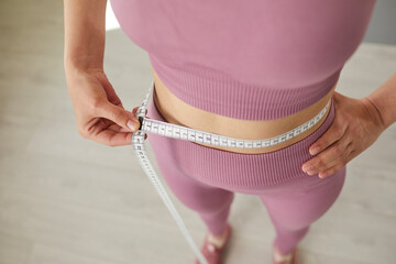 Fototapeta premium Cropped photo of faceless unrecognizable slim sporty woman wearing sportswear measure the waist with measuring tape standing on the floor at home. Slimming, weight control concept.