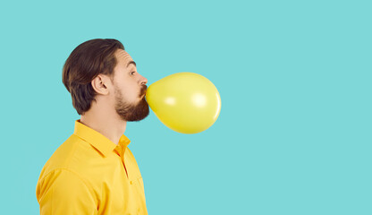 Young bearded man with funny expression inflates yellow ball on turquoise background. Portrait in...