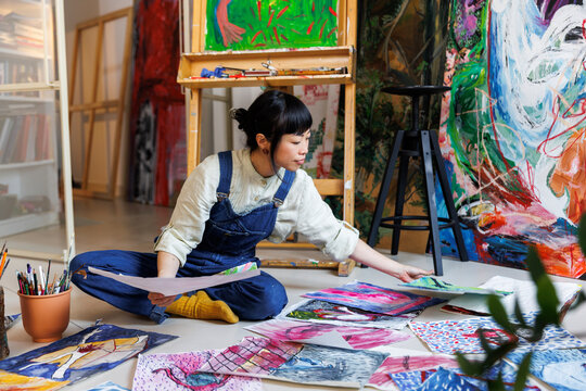 Female artist arranging various paintings on floor at studio