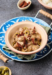 A hot dish, stewed beef with dried tomatoes and olives in a clay bowl on a blue meallic tray on a dark concrete background. Beef recipes.