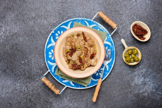 A hot dish, stewed beef with dried tomatoes and olives in a clay bowl on a blue meallic tray on a dark concrete background. Beef recipes.