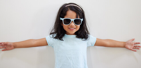 Girl, child and smile with sunglasses by wall for summer, holiday and playful at family home. Kid,...
