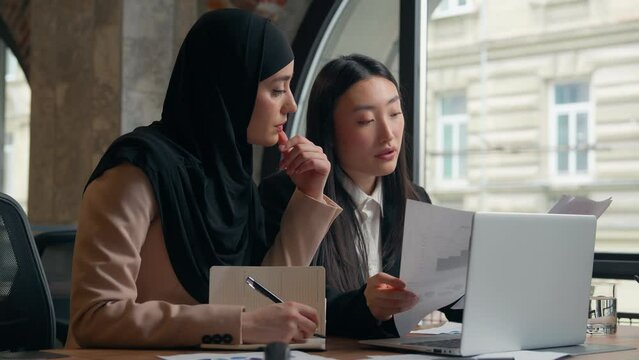 Two business women student girls multiracial female multiethnic businesswomen Asian Korean boss mentor woman teach help Muslim Islamic intern in hijab colleagues discuss documents teamwork in office