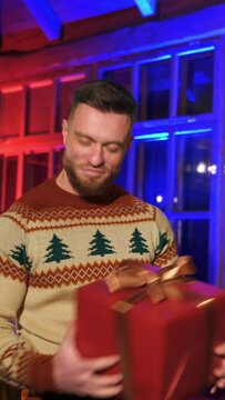 Smiling man offering new year present. Portrait of a handsome man in knitted Christmas sweater holding a box with gift on Christmas decorated room background. Vertical video