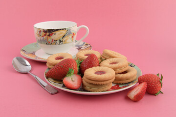 a plate of strawberry cream cookies and strawberries and a colorful tea cup on a pink background