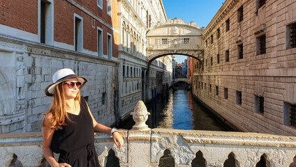 Tourist woman with had and sunglasses under the Bridge of Sighs in Venice, Veneto, Northern Italy, Europe. Last thing criminals saw before the went to jail. Historical building. Rio di Palazzo - obrazy, fototapety, plakaty