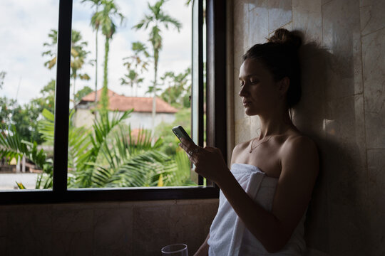 Woman in Towel Scrolling on Her Phone Near Opened Window