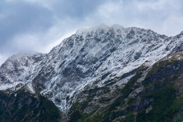 Photograph of cloud covered snow capped mountains with lush foliage in Fiordland National Park on the South Island of New Zealand