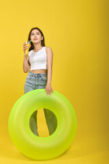 Asian young brunette woman with glass fresh juice dreamily enjoying drink while standing with large inflatable circle. Cute long-haired girl on a yellow background posing with an inflatable circle
