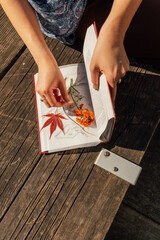 woman pressing freshly picked flowers and leaves in book on outdoor bench