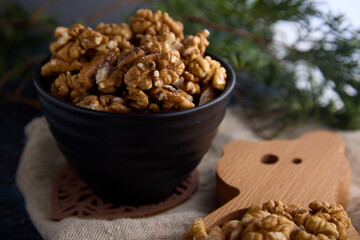 On a blurred background, on an old burlap, there is a black bowl filled with halves of delicious walnuts. Lots of peeled walnuts in a ceramic black bowl on wooden stands on the table