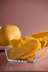  In a transparent glass dish, dry delicious mango slices and a whole mango are laid out on a plate. Vertical photo of mango fruits beautifully laid out in a glass dish on a pink background.